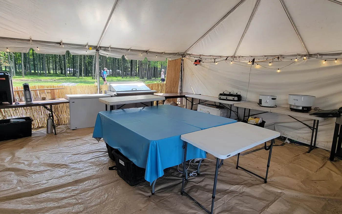 Inside the mobile kitchen tent: prep tables, cooktops, a griddle and coolers, with woods behind